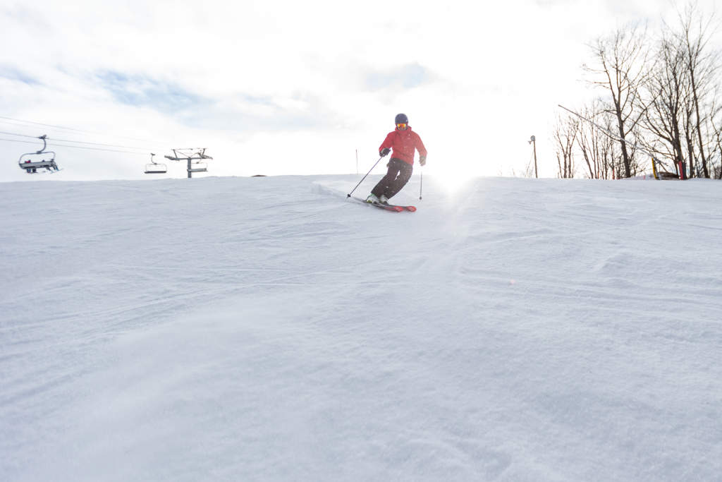 Male skier in a red jacket carving a turn at the top of the ski hill with the chairlift in the background